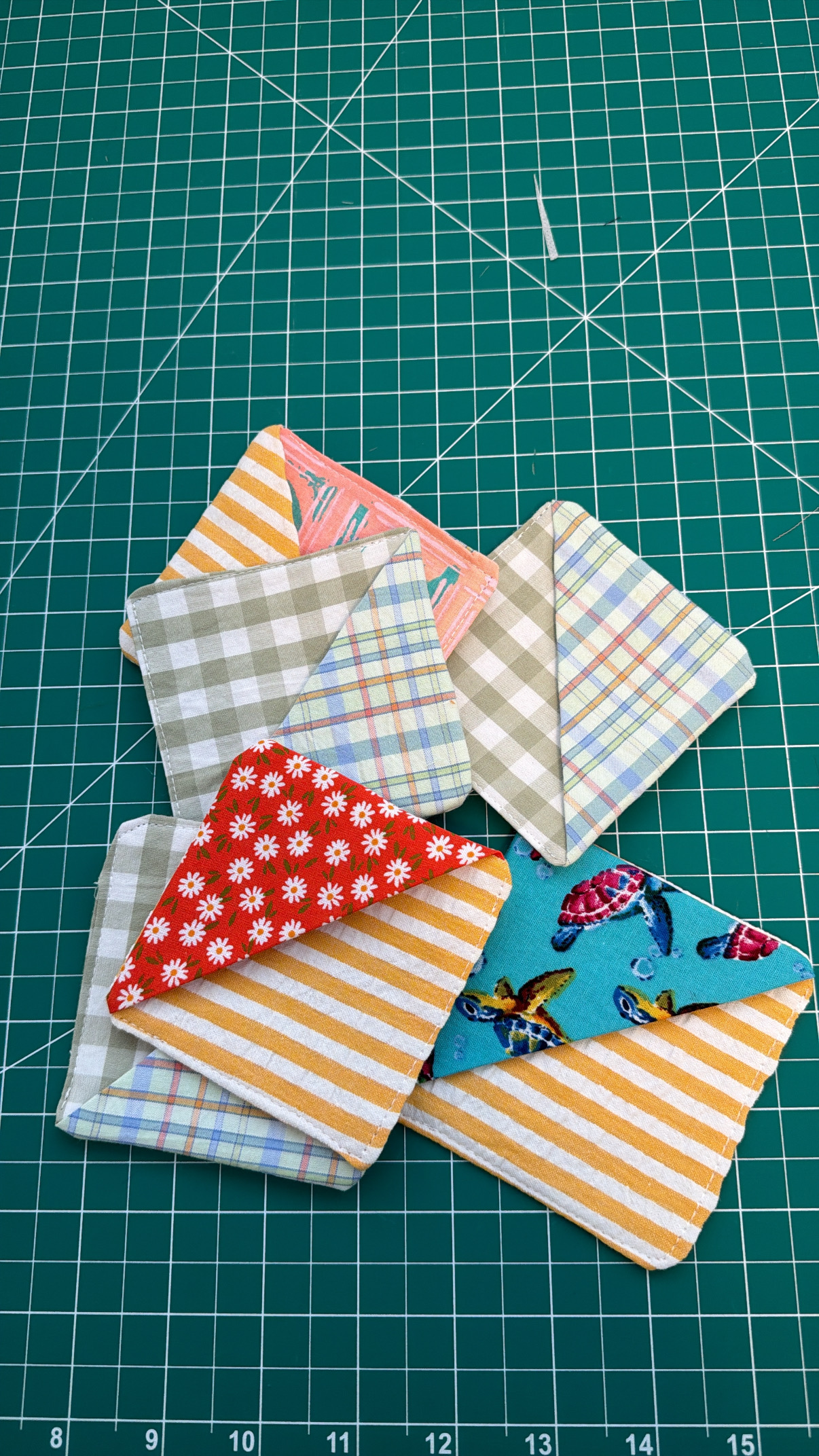 A pile of finished fabric corner bookmarks in a mix of scrap fabrics, florals, plaids, gingham, and a turtle print
